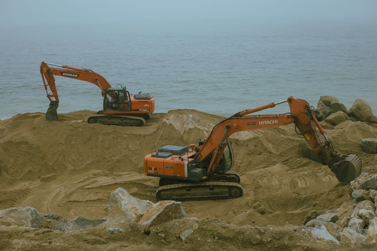 who-we-are Two excavators operate at a sandy construction site by the sea, moving sand and rock.